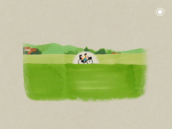 Screenshot from Five Years Old Memories of a parent on a bicycle with a child in the backseat, seen riding along a grassy ridge from a distance. There are hills and houses in the background
